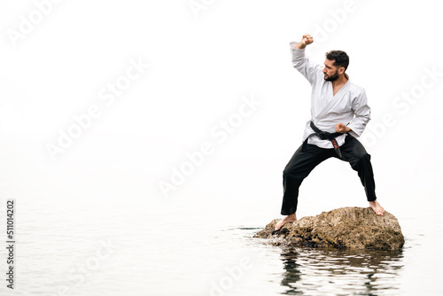 man standing on a rock in the sea wearing a karate kimono in a defensive position with the word "Bushido" on his belt and a completely white background.
