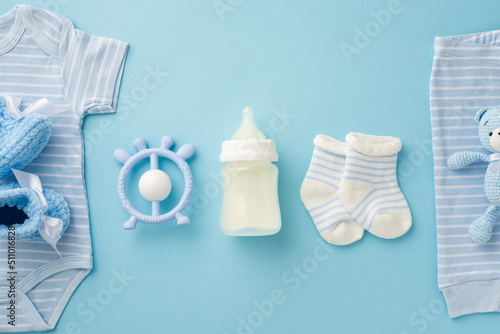 Baby accessories concept. Top view photo of blue infant clothes bodysuit panties knitted booties socks teddy bear toy rattle and milk bottle on isolated pastel blue background