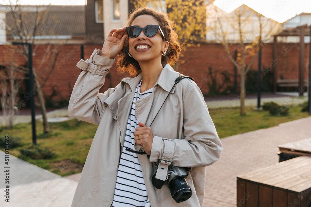 Fototapeta premium Positive young african woman admiring the autumn sky looking at it through sunglasses. Brunette with curly hair wears casual clothes. Lifestyle concept