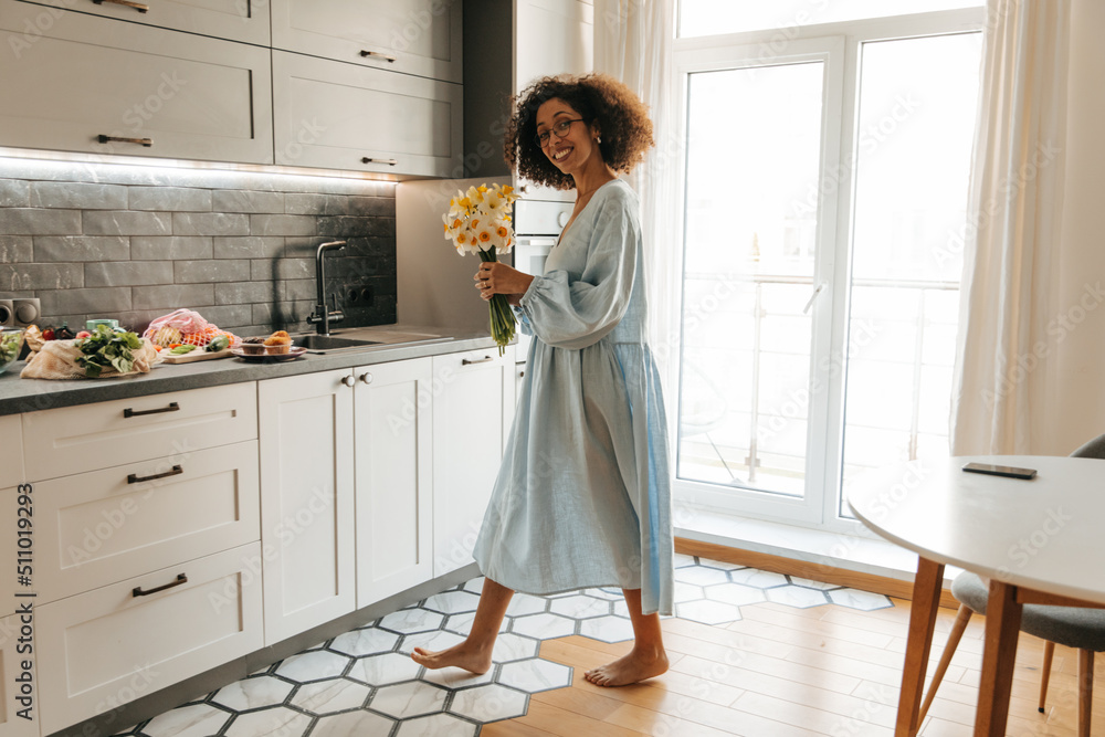 Full length beautiful young african woman looking at camera, holding flowers standing in kitchen interior. Brunette with afro hair wears blue dress at home. Mood, lifestyle, concept. Full length beautiful young african woman looking at camera, holding flowers standing in kitchen interior. Brunette with afro hair wears blue dress at home. Mood, lifestyle, concept.