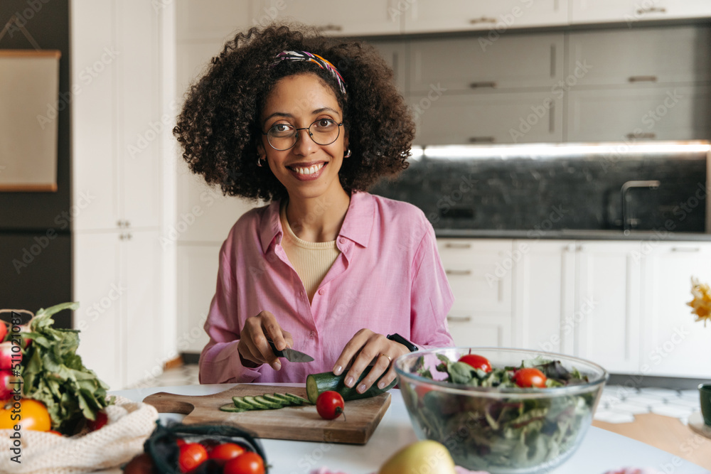 Positive young african brunette woman preparing healthy meal from vegetables sitting in kitchen. Girl wears glasses, pink shirt at home. Vegan salad concept Positive young african brunette woman preparing healthy meal from vegetables sitting in kitchen. Girl wears glasses, pink shirt at home. Vegan salad concept