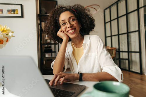 Happy young african woman looking at camera sitting at laptop home. Brunette with curly hair wears white shirt. Mood, lifestyle, concept. photo