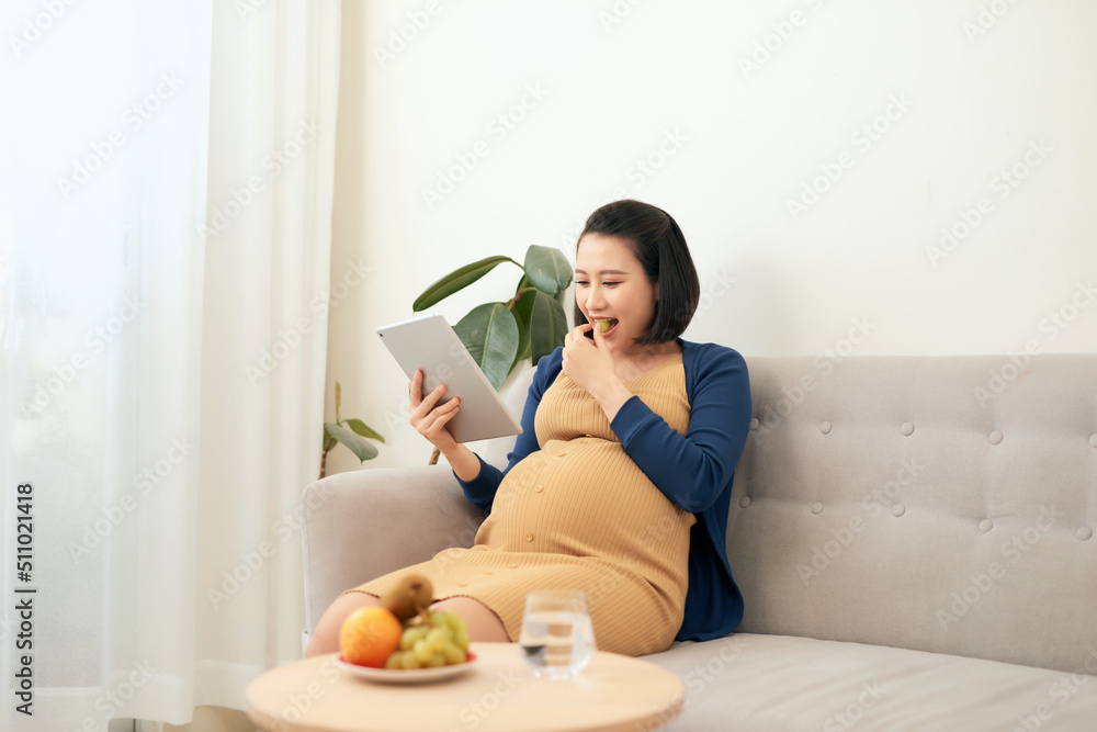 Beautiful young Asian pregnant woman using tablet and eating fruit when sitting on sofa. Beautiful young Asian pregnant woman using tablet and eating fruit when sitting on sofa.