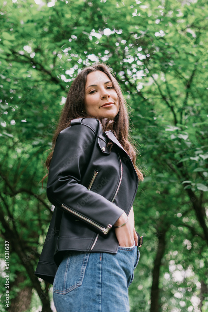 Portrait of a young woman. Leather jacket and jeans, the model poses against a background of green trees. Summer mood Portrait of a young woman. Leather jacket and jeans, the model poses against a background of green trees. Summer mood
