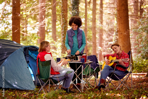 Group Of Female Friends On Camping Holiday In Forest Eating Meal And Singing Along To Guitar