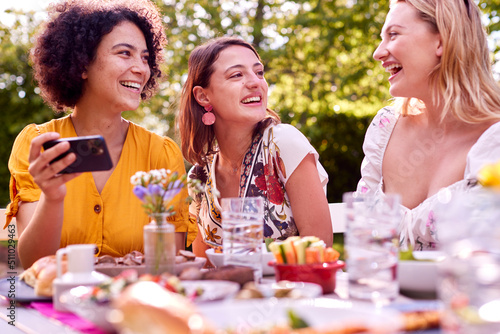 Three Female Friends Looking At Photos On Mobile Phone Eating Meal Outdoors In Summer Garden At Home