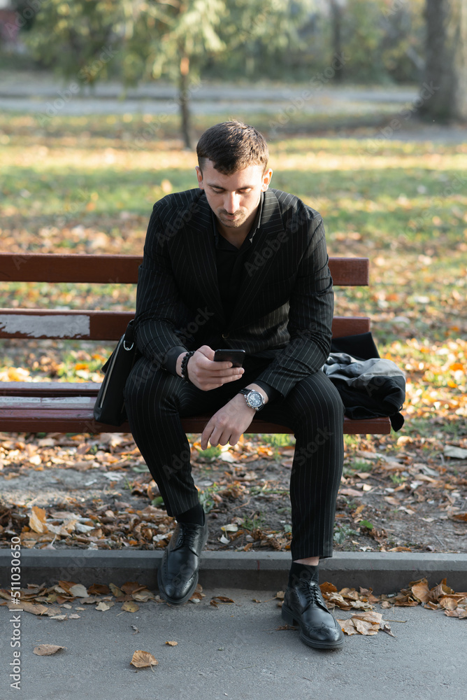 Obraz premium A young businessman is sitting on a park bench using a smartphone