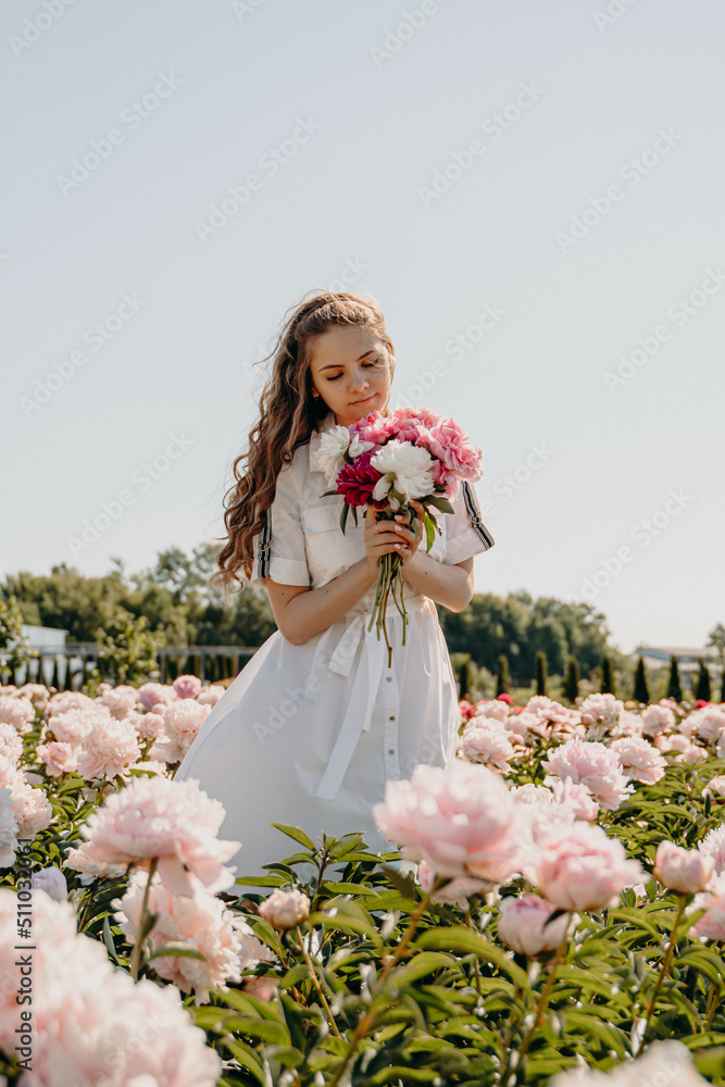 Happy young sexy girl standing in the middle of a flower field holding a peony bouquet. Single woman in white dress resting on pink background enjoying summer day. Happy young sexy girl standing in the middle of a flower field holding a peony bouquet. Single woman in white dress resting on pink background enjoying summer day.