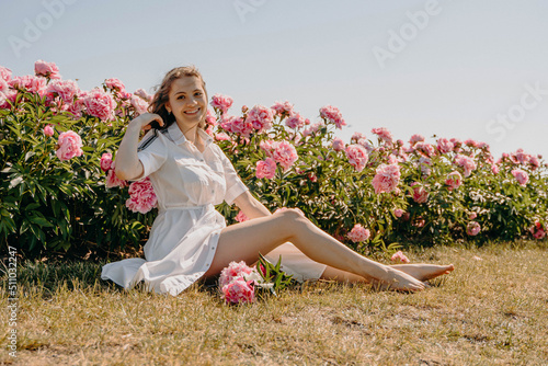 Beautiful sexy young girl sitting in a flower garden, young woman in a field of peonies. The girl is smiling, dressed in a white dress with beautiful legs. The concept of freedom, recreation. Sunny su