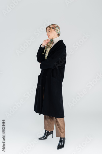full length of woman in kerchief and black coat touching chin on grey background.