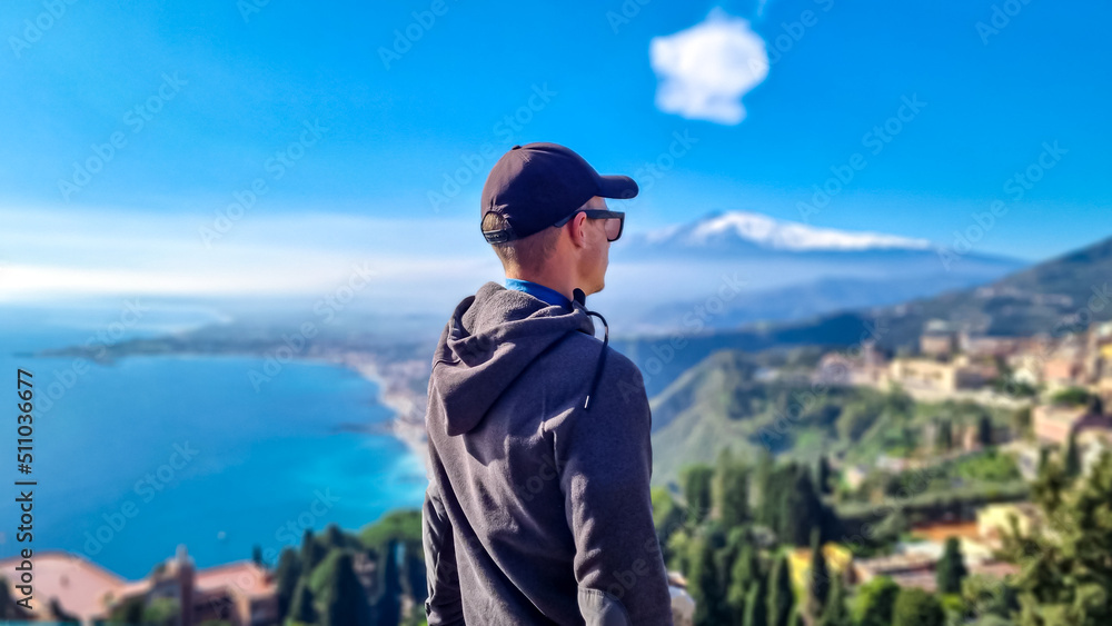 Naklejka premium Tourist man with panoramic view on snow capped Mount Etna volcano and the Mediterranean coastline on a sunny day seen from ancient Greek theater of Taormina, island Sicily, Italy, Europe, EU. Awe