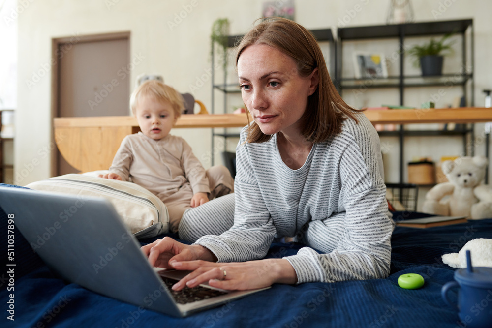 Young woman lying on bed and typing on laptop, she doing online work having a little child Young woman lying on bed and typing on laptop, she doing online work having a little child