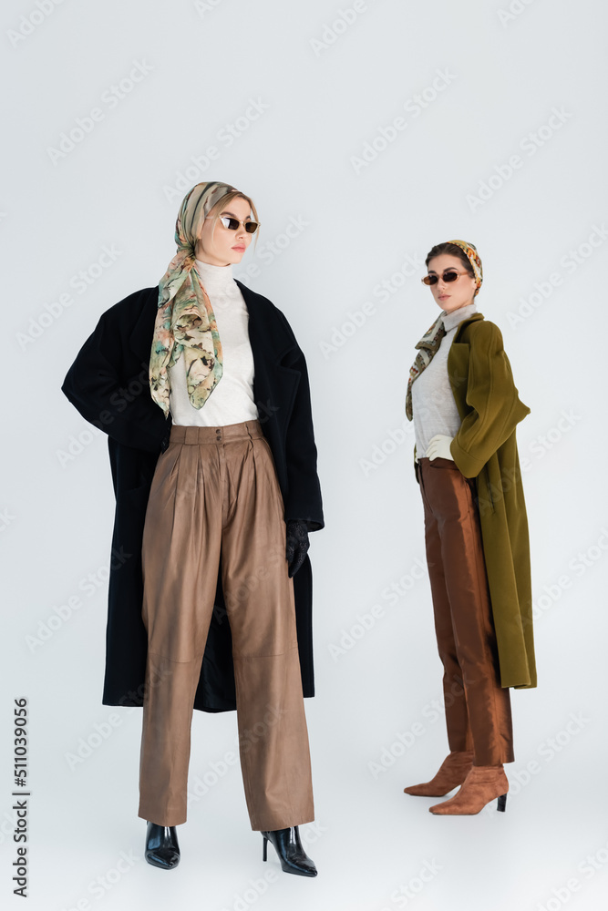 full length of elegant women in coats and sunglasses posing on grey background.