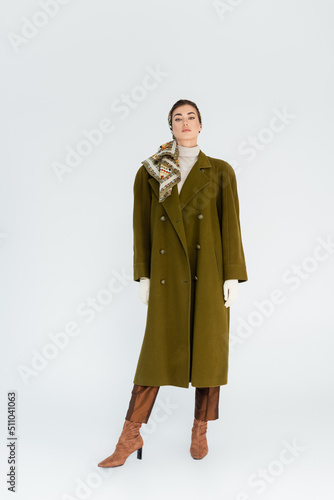 full length of woman in green stylish coat and headscarf looking at camera on white background.