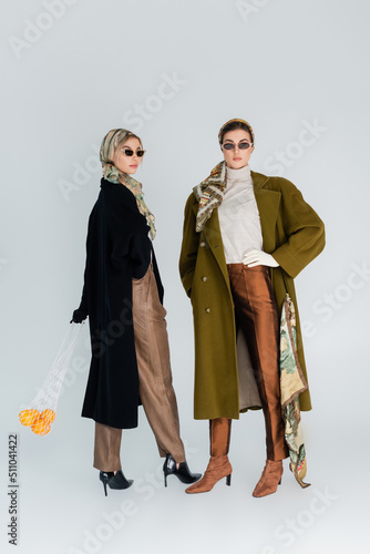 full length of trendy woman with oranges in mesh bag near friend standing with hand on waist on grey background.