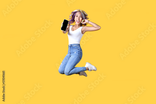Fototapeta Naklejka Na Ścianę i Meble -  Full length portrait of young Asian woman holding smartphone jumping on yellow background. Cheerful young female jumping up and showing mobile phone with empty screen, in studio