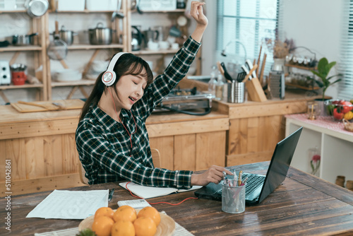 asian female student is relaxing at dining table listening to upbeat songs on laptop. chinese girl taking break from preparing for test is enjoying online streaming music with hand raising in the air