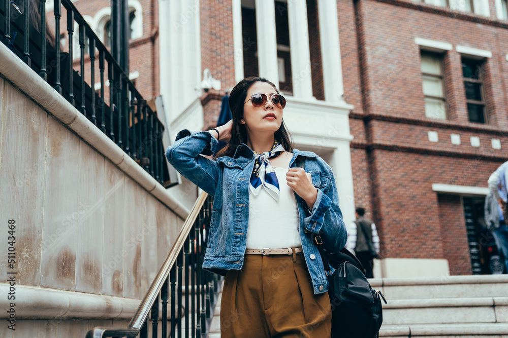 Fototapeta premium stylish chinese lady hand combing hair is standing on outdoor staircase with metal handrail. modern millennial woman is looking into distance from a high place, background red brick building