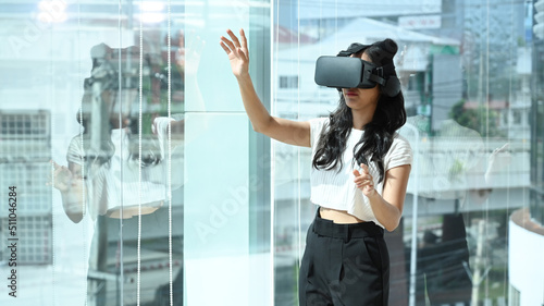 Excited businesswoman wearing virtual reality glasses, interacting with digital interface while standing near in large window at modern office.
