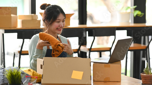 Pretty asian woman showing clothes front of laptop computer, present product via live streaming online. E-Commerce, online sales concept