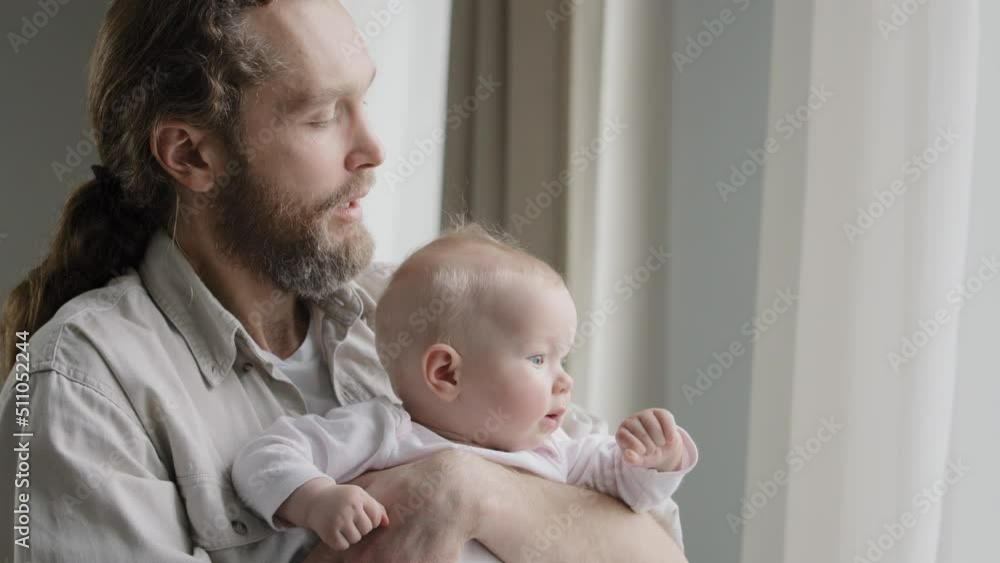 Close-up caucasian father bearded daddy holding baby daughter son infant newborn looking out window with curtains dad blowing to short blonde hair of kid child playing at home. Fatherhood protection Close-up caucasian father bearded daddy holding baby daughter son infant newborn looking out window with curtains dad blowing to short blonde hair of kid child playing at home. Fatherhood protection