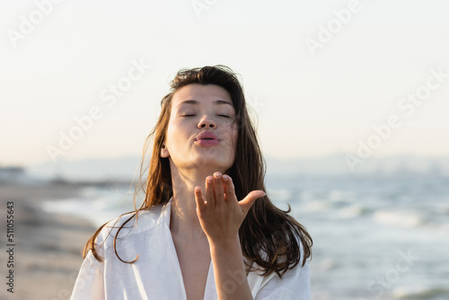 Brunette woman in white shirt blowing air kiss on blurred beach.