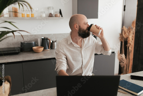 A man with a beard works remotely in his modern kitchen using a laptop and drinks a hot drink