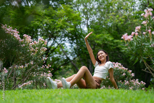 Portrait photo of a young beautiful asian female lady in white top sitting on the grass embracing the breeze wind and soft sunlight relaxingly and chillingly in a public park © asean studio