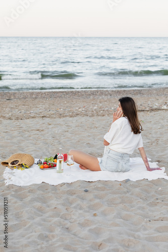 Woman talking on smartphone near wine and fruits on sea coast. photo