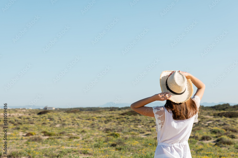 Back view of young woman holding straw hat near grass on beach at background.