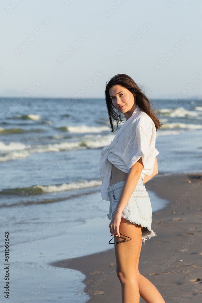Pretty brunette woman in shirt holding sunglasses and looking at camera on beach.