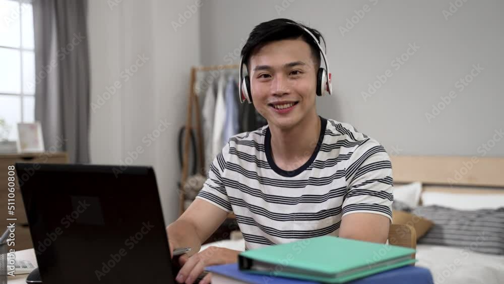 portrait of an asian young male wearing headset is turning to look at the camera with a smile while learning from home on laptop in the bedroom during lockdown portrait of an asian young male wearing headset is turning to look at the camera with a smile while learning from home on laptop in the bedroom during lockdown