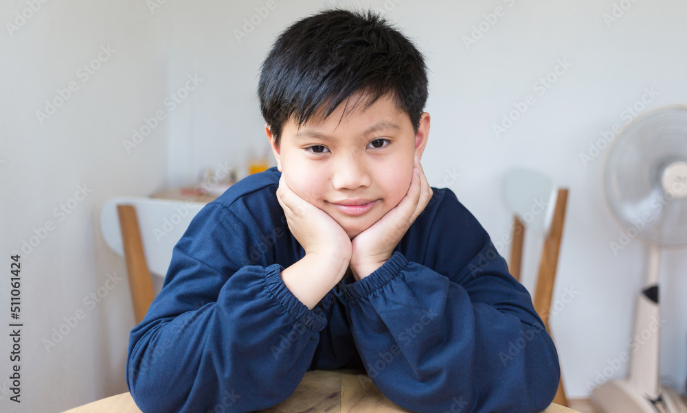 Cute Asian boy with white skin smiling looking in camera happily resting chins on hands-on wooden table. Close-up portrait of a cute child. Cute Asian boy with white skin smiling looking in camera happily resting chins on hands-on wooden table. Close-up portrait of a cute child.