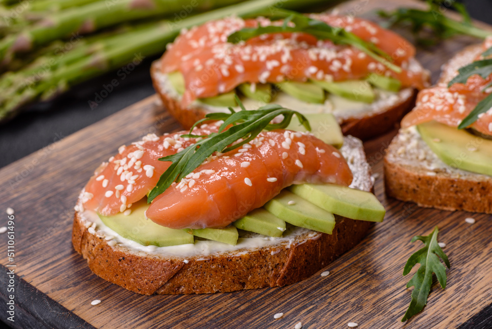 Toast sandwich with butter, avocado and salmon, decorated with arugula and sesame seeds, on a black stone background Toast sandwich with butter, avocado and salmon, decorated with arugula and sesame seeds, on a black stone background