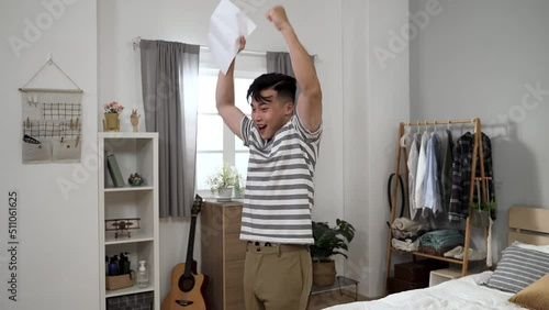 happy asian young man looking at paper document in surprise is kissing it with raised fists while celebrating success of getting admitted to dream school at home