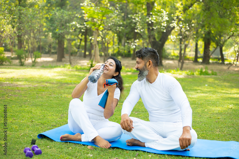 Indian man and woman sitting in the park drinking water after yoga exercise workout, Asian couple relaxing in the garden together outdoor.