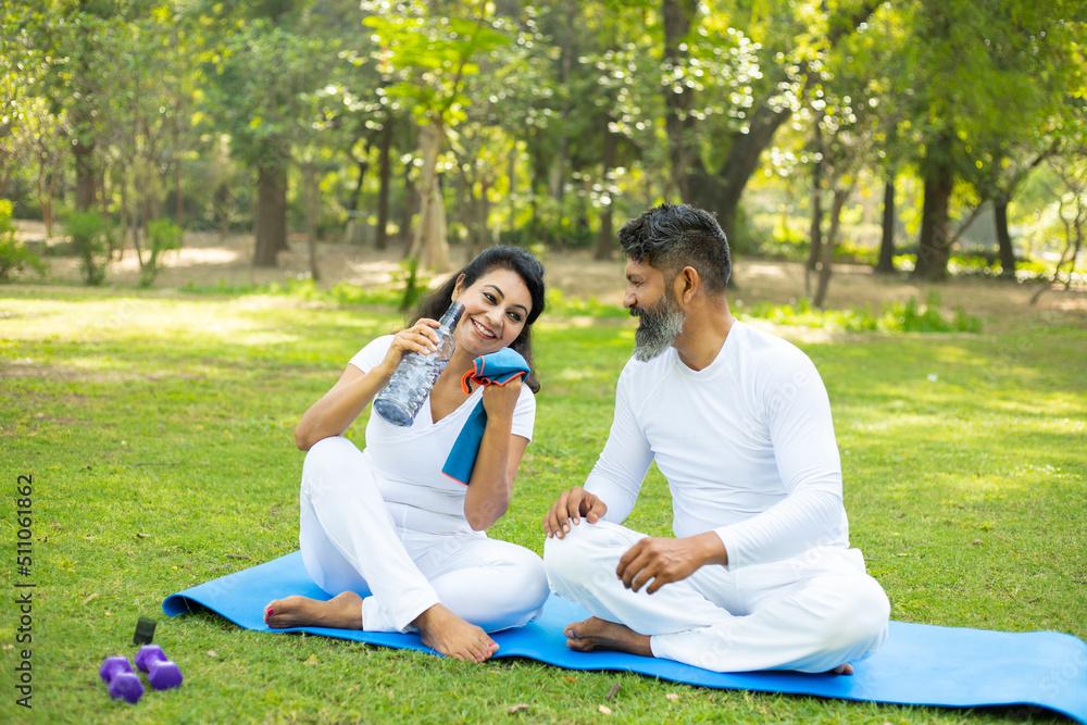 Indian man and woman sitting in the park drinking water after yoga exercise workout, Asian couple relaxing in the garden together outdoor. Indian man and woman sitting in the park drinking water after yoga exercise workout, Asian couple relaxing in the garden together outdoor.