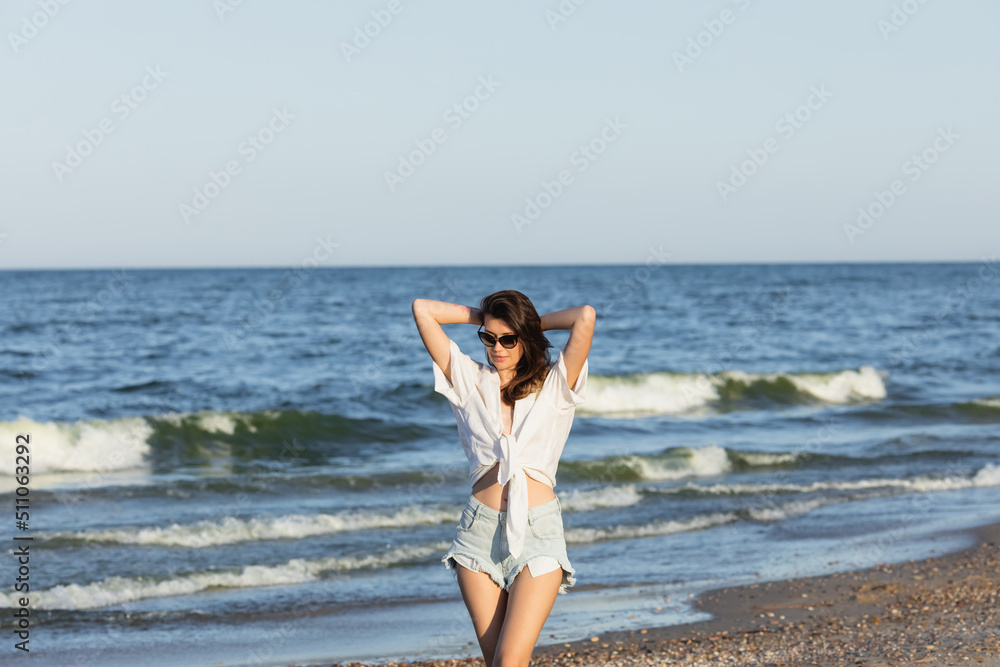 Young woman in sunglasses and denim shorts standing near blurred sea.