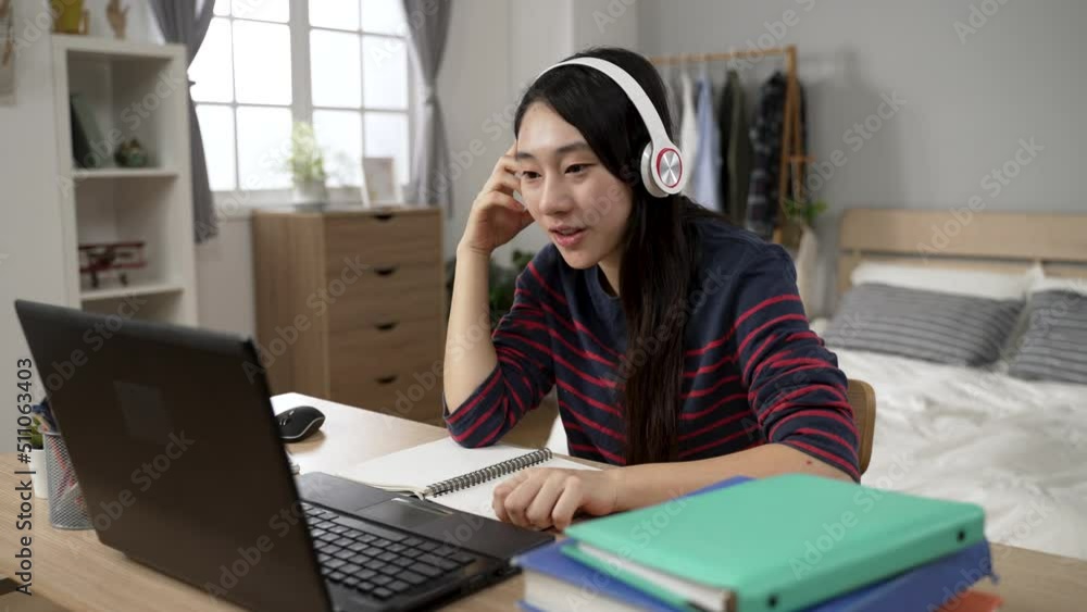 happy asian college girl learning from home with earphone is waving hi to laptop screen while having online virtual class in the bedroom during daytime. happy asian college girl learning from home with earphone is waving hi to laptop screen while having online virtual class in the bedroom during daytime.