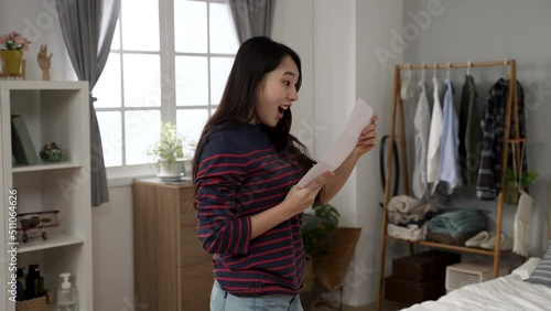 excited Japanese female unfolding letter with job offer message while walking in bedroom is spinning in place with clenched fist and reading details with smile