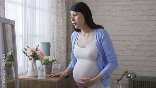 Картината върху платно asian expectant female standing by the cupboard is holding her tummy and groaning in pain due to labor contraction at bright home interior