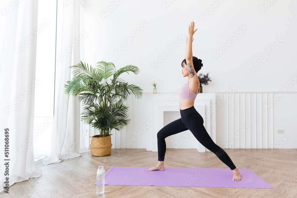 Young smiling attractive sporty Asian woman practicing yoga, doing Virabhadrasana 1 exercise, meditating in Warrior One yoga pose, indoor working out at home, wearing sportswear. Full length photo.