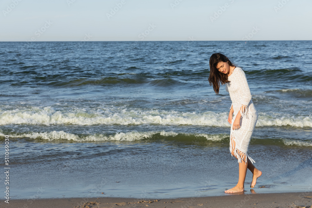 Full length of brunette woman in dress walking on sand near sea.