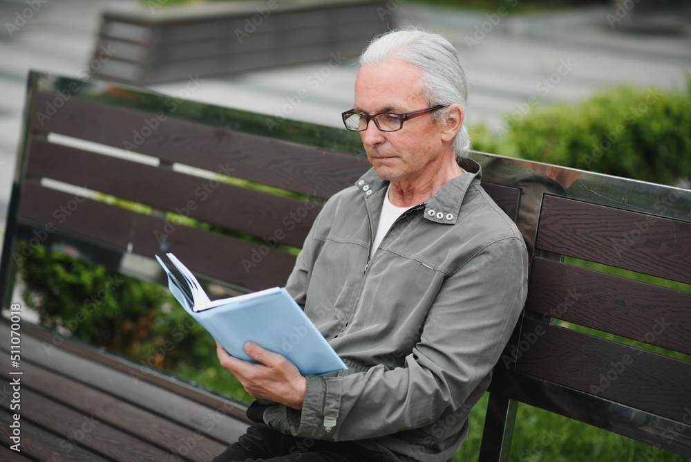 Fototapeta premium Portrait of senior man reading on bench during summer day.