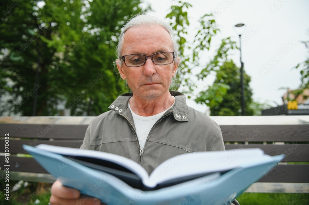 Old man with gray hair reads a book on a bench in the park. Rest in the park. Old man with gray hair reads a book on a bench in the park. Rest in the park.
