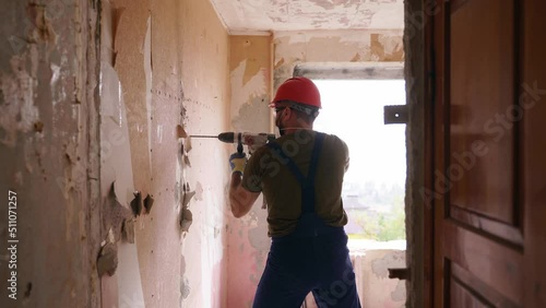 Builder working with rotary hammer drill. Contractor drills holes in concrete with jackhammer to demolish the wall for apartment remodelling and renovation. Electrician in uniform installs wiring.