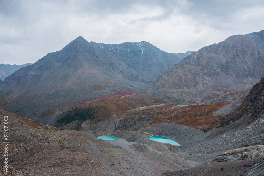Awesome aerial view to turquoise eyes-lakes and pyramid shaped high mountain under dramatic gray cloudy sky. Two beautiful mountain lakes among fading autumn colors in overcast. Small glacial lakes.
