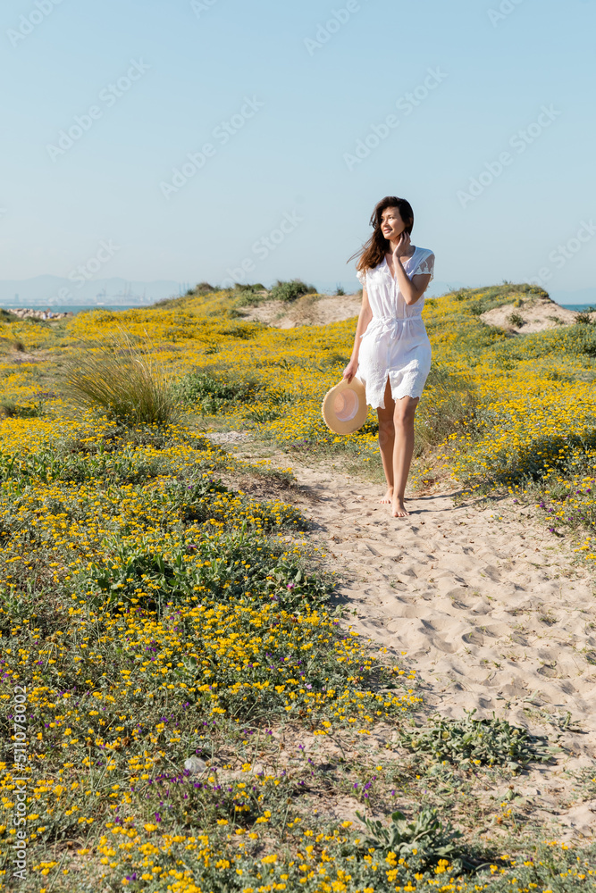 Pretty woman holding straw hat while standing near flowers on beach.