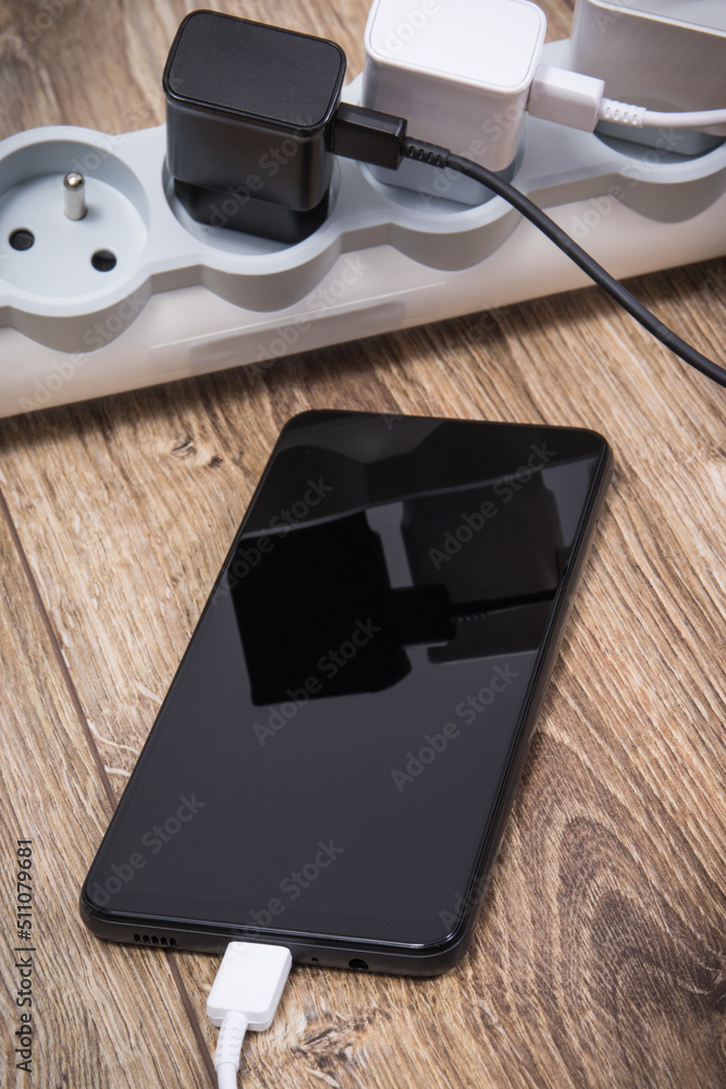 Black smartphone and chargers connected to electrical power strip. Various devices charging concept Black smartphone and chargers connected to electrical power strip. Various devices charging concept