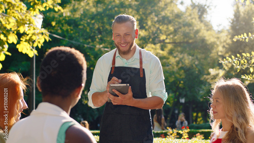 A very good looking waiter with his hair slicked back, is taking down orders in his notebook, he s talking to three women taking their orders and discussing while being in a park scenery